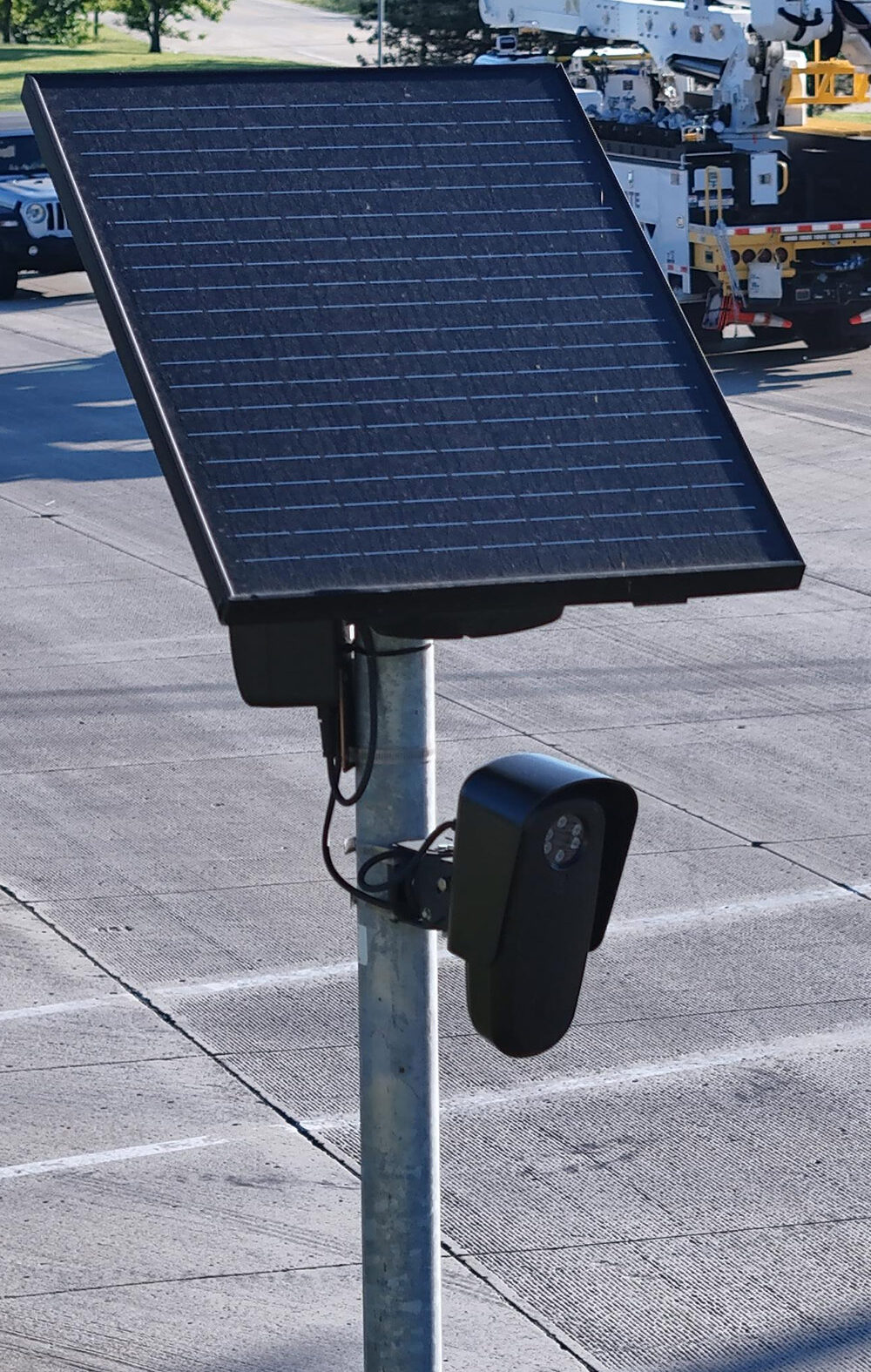 A solar-powered Flock camera is mounted on a pole beside a busy multi-lane road, with cars and a utility truck driving through the intersection in the background.