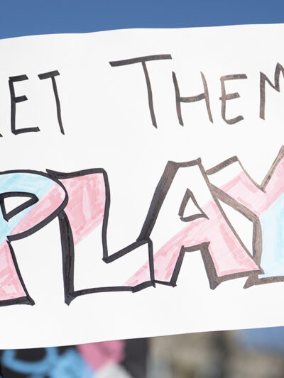 A hand holds a handmade sign reading "LET THEM PLAY," with the word "PLAY" colored in pink, blue, and white transgender pride colors, during a rally under a clear blue sky.
