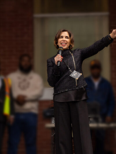 Yasmin Cader stands on an outdoor stage in front of a brick building, speaking into a microphone and gesturing with papers in her raised hand, while several people stand blurred in the background.