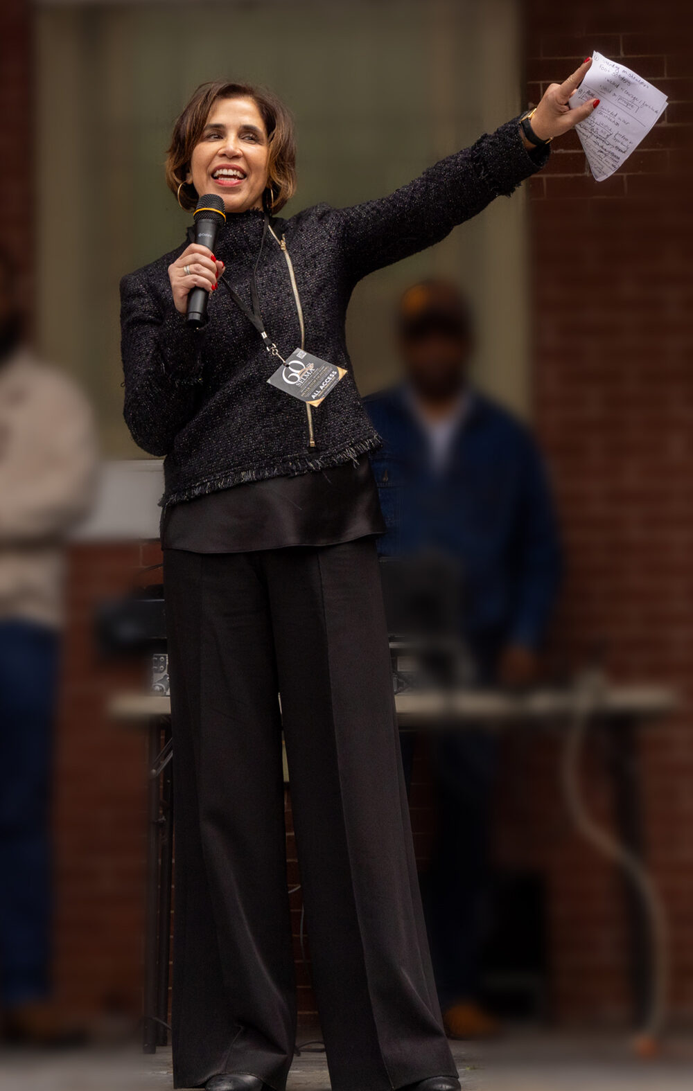 Yasmin Cader stands on an outdoor stage in front of a brick building, speaking into a microphone and gesturing with papers in her raised hand, while several people stand blurred in the background.