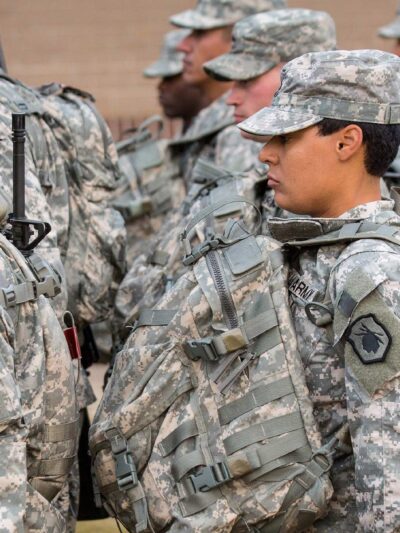 U.S. Army soldiers, including women in the foreground, stand in formation outdoors wearing digital camouflage uniforms, patrol caps, and large tactical backpacks with straps and attached gear, all facing the same direction.