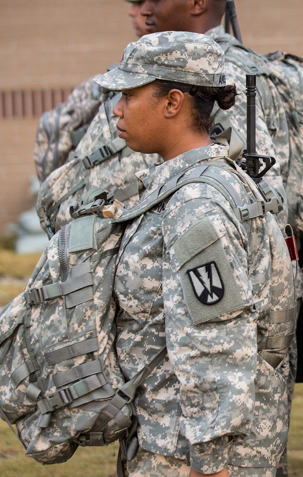U.S. Army soldiers, including women in the foreground, stand in formation outdoors wearing digital camouflage uniforms, patrol caps, and large tactical backpacks with straps and attached gear, all facing the same direction.