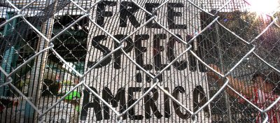 A sign reading 'Free Speech in America' is visible through a chainlink fence