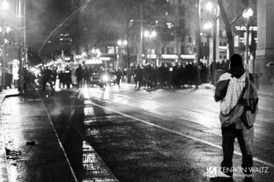 A protestor stands in the foreground, facing a line of police in riot gear about 10 yards away
