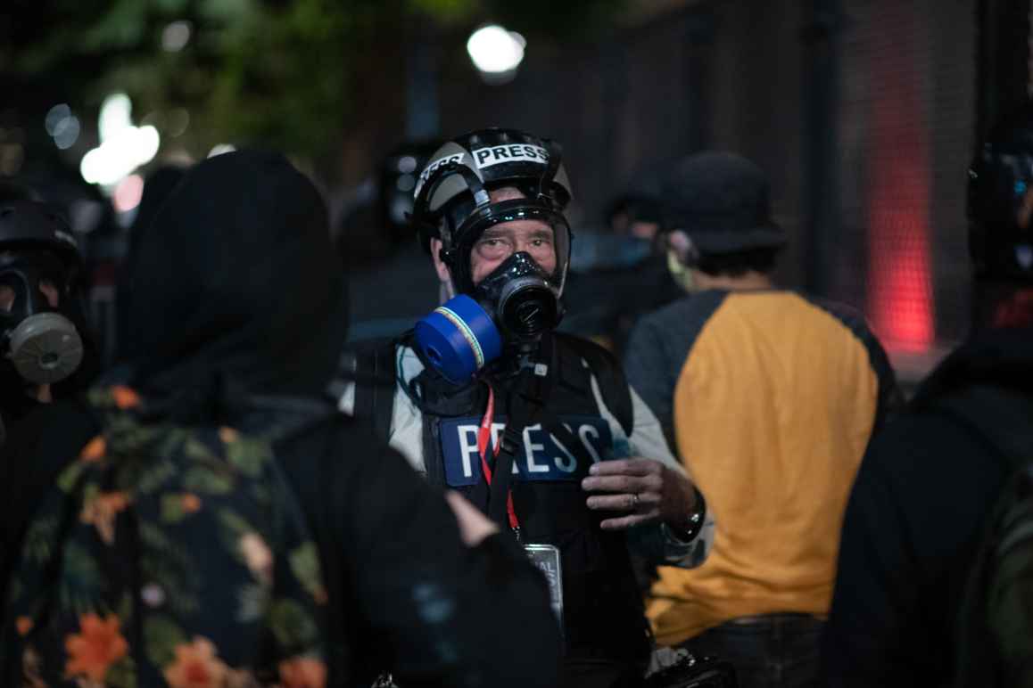 A journalist, wearing a helmet and gas mask, is standing in a crowd at a protest