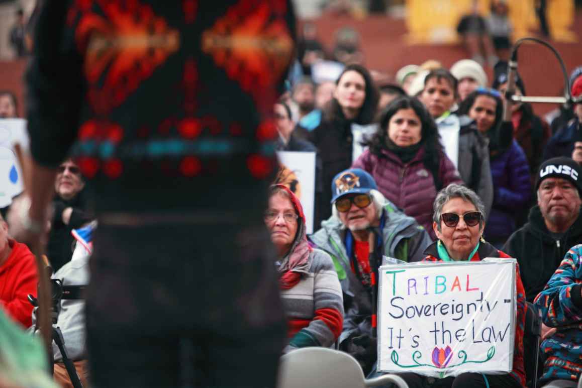 A crowd of people listens to a man speak. A woman holds a sign that reads 'Tribal Sovereignty, It's the law'