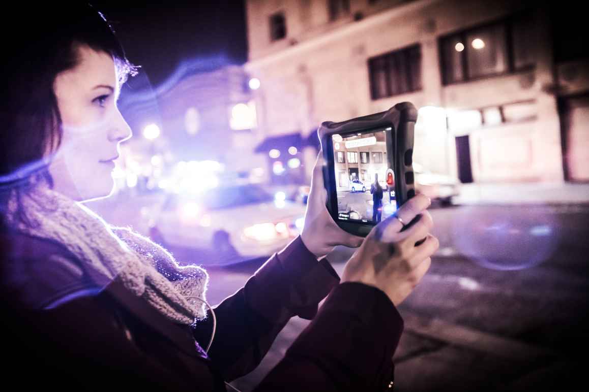 Carrie Medina holds a tablet to film the police on the street. Police lights blur into the foreground.