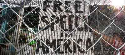 A sign reading 'Free Speech in America' is visible through a chainlink fence