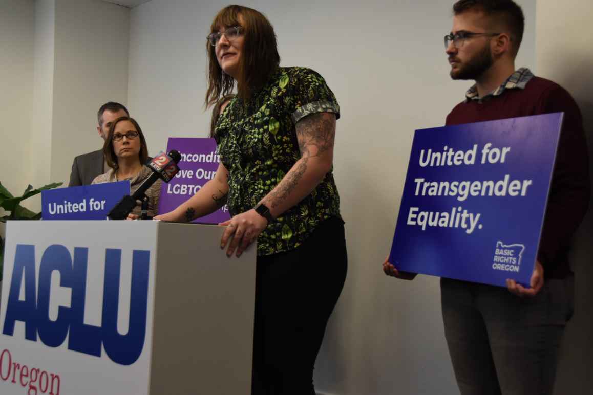 Brook Shelley stands at podium, people are holding signs in the background that have messages supporting transgender youth 