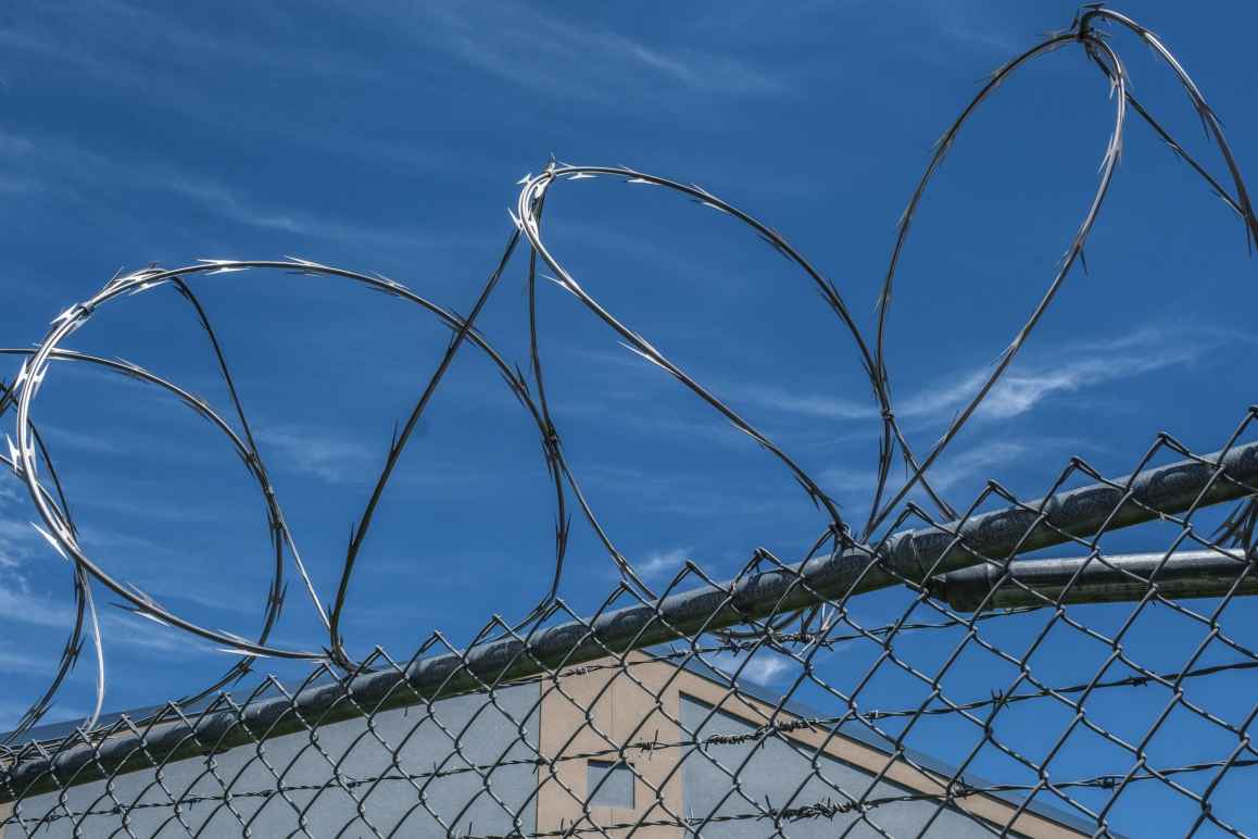 Close up image of a prison fence with barbed wire at the top glinting in the sun