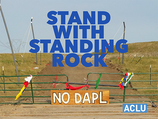 A closed metal gate stands closed with a sign reading 'No DAPL' in front of it. Text reads 'Stand with Standing Rock'
