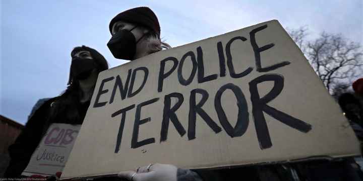 A demonstrator wearing a black hospital mask and black beanie hat holds up a cardboard sign that reads, "END POLICE TERROR".