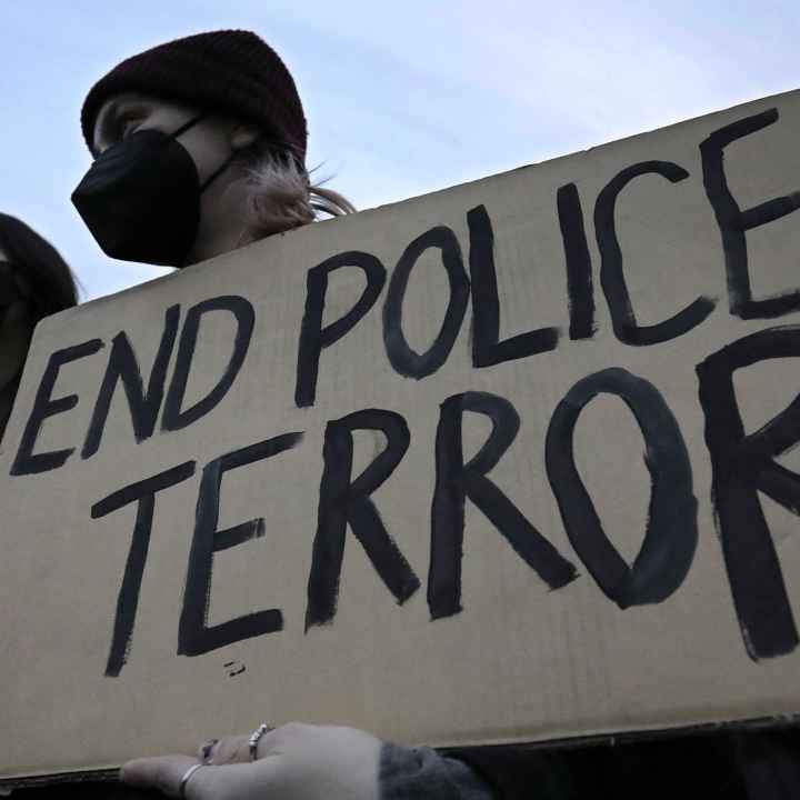 A demonstrator wearing a black hospital mask and black beanie hat holds up a cardboard sign that reads, "END POLICE TERROR".