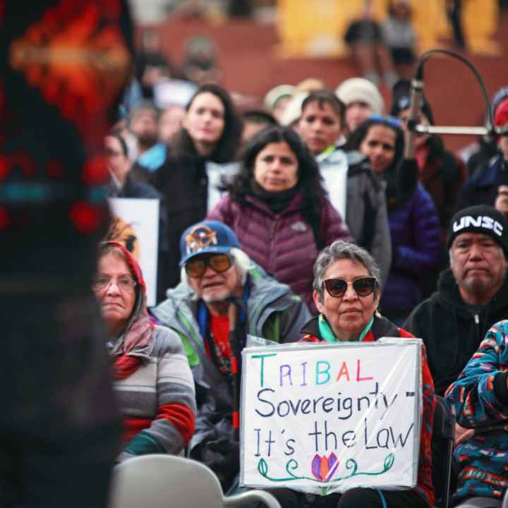 A crowd of people listens to a man speak. A woman holds a sign that reads 'Tribal Sovereignty, It's the law'