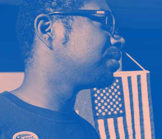 A man wears an "I Voted" stick with the American flag hanging in the background. Image tinted blue and pink.